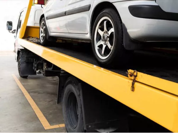 A car transportation company drives a silver car on a yellow auto transport trailer.