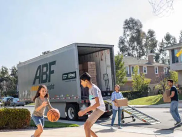 Two kids playing basketball in front of a U-Pack trailer while parents unload moving boxes.