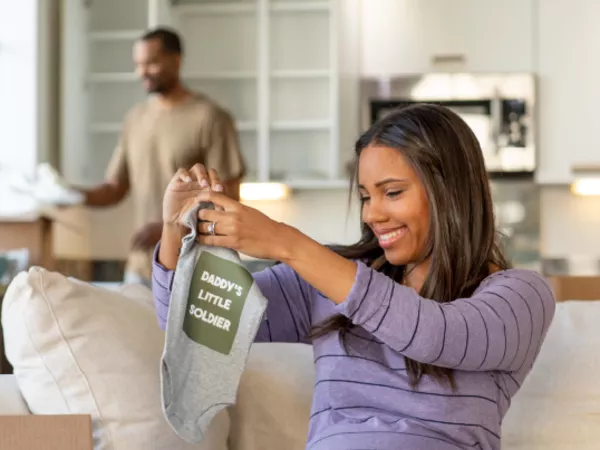 An expecting mother holds up baby clothes that reads Daddy's Little Soldier